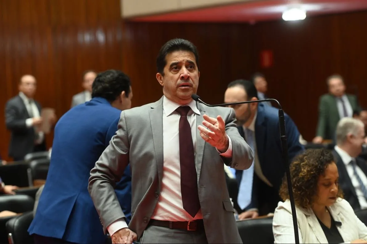 Deputado Sargento Rodrigues fala ao microfone em plenário, vestindo terno cinza e gravata vinho, com expressão firme e gesto com a mão direita. Ao fundo, outros parlamentares conversam e acompanham a sessão legislativa.