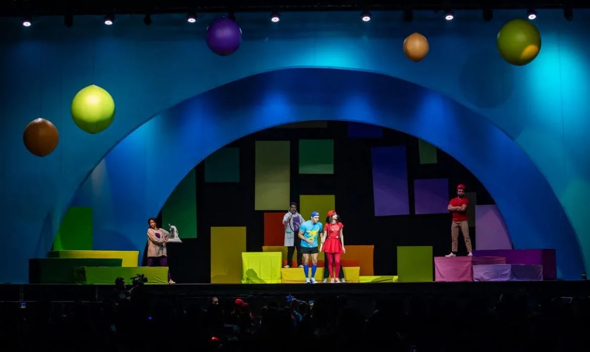 Cenário colorido do espetáculo infantil “Luccas e Gi em: O Mundo de Magia e Fantasia”, com Luccas Neto, Gi e elenco em cena no palco do Be Fly Hall, em Belo Horizonte, sob iluminação teatral vibrante e elementos cenográficos lúdicos.