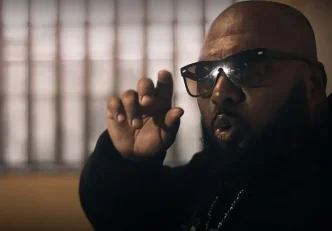Close marcante de Black W durante as gravações do clipe “Lágrimas de Um Detento” — o rapper aparece com expressão intensa, óculos escuros e gesto enfático com a mão, em um ambiente de iluminação dramática, reforçando a atmosfera de denúncia social e vivência carcerária que permeia a canção.
