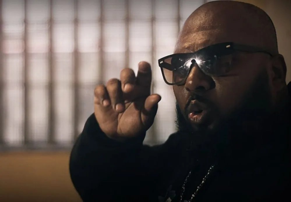 Close marcante de Black W durante as gravações do clipe “Lágrimas de Um Detento” — o rapper aparece com expressão intensa, óculos escuros e gesto enfático com a mão, em um ambiente de iluminação dramática, reforçando a atmosfera de denúncia social e vivência carcerária que permeia a canção.