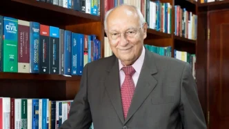 Retrato do professor e jurista Ives Gandra da Silva Martins em sua biblioteca pessoal, vestindo terno cinza escuro e gravata vermelha, cercado por estantes repletas de livros jurídicos, com destaque para obras de direito civil e constitucional ao fundo.