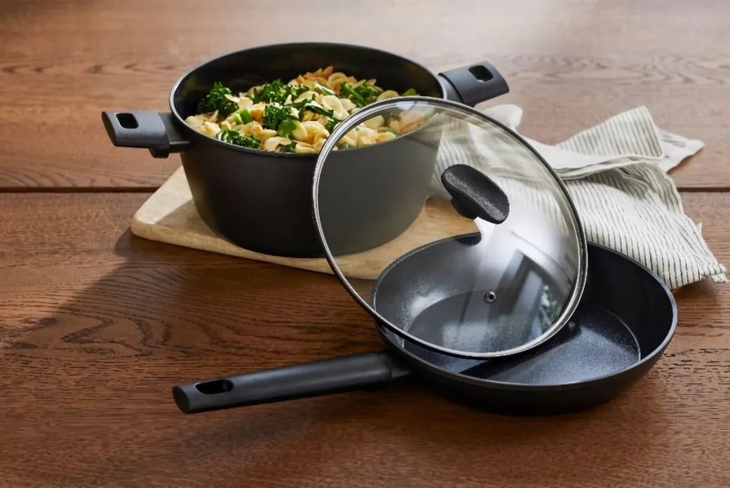 Panelas da marca francesa Cuisinox sobre mesa de madeira, destacando a caçarola com preparo de massa e vegetais e uma frigideira com tampa de vidro, itens oferecidos na campanha de fidelidade do Supernosso em Belo Horizonte.
