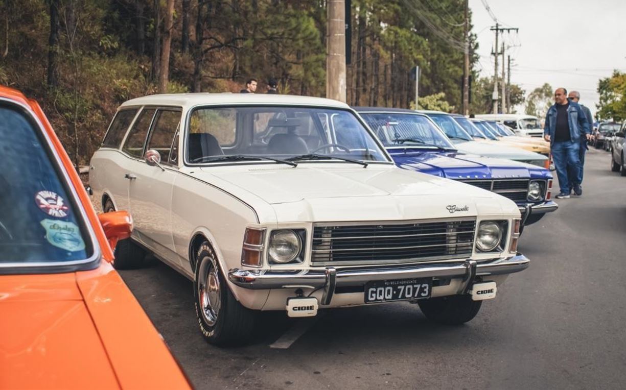 Linha de veículos clássicos estacionados em Belo Horizonte durante encontro automotivo, com destaque para um Chevrolet Caravan branco.