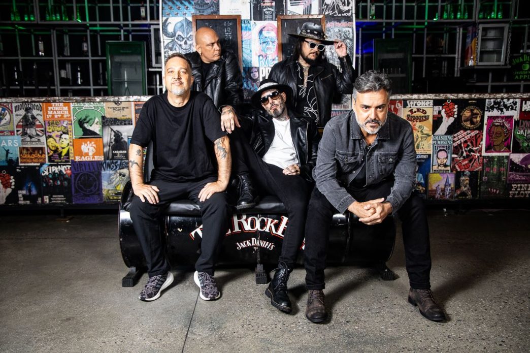Integrantes da banda Detonautas posam com atitude em ambiente underground, cercados por pôsteres de ícones do rock mundial.