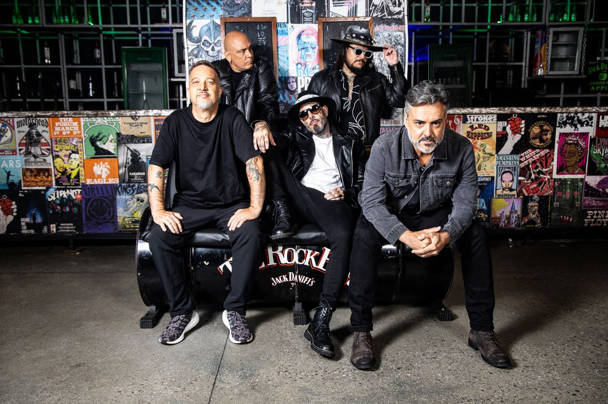 Integrantes da banda Detonautas posam com atitude em ambiente underground, cercados por pôsteres de ícones do rock mundial.