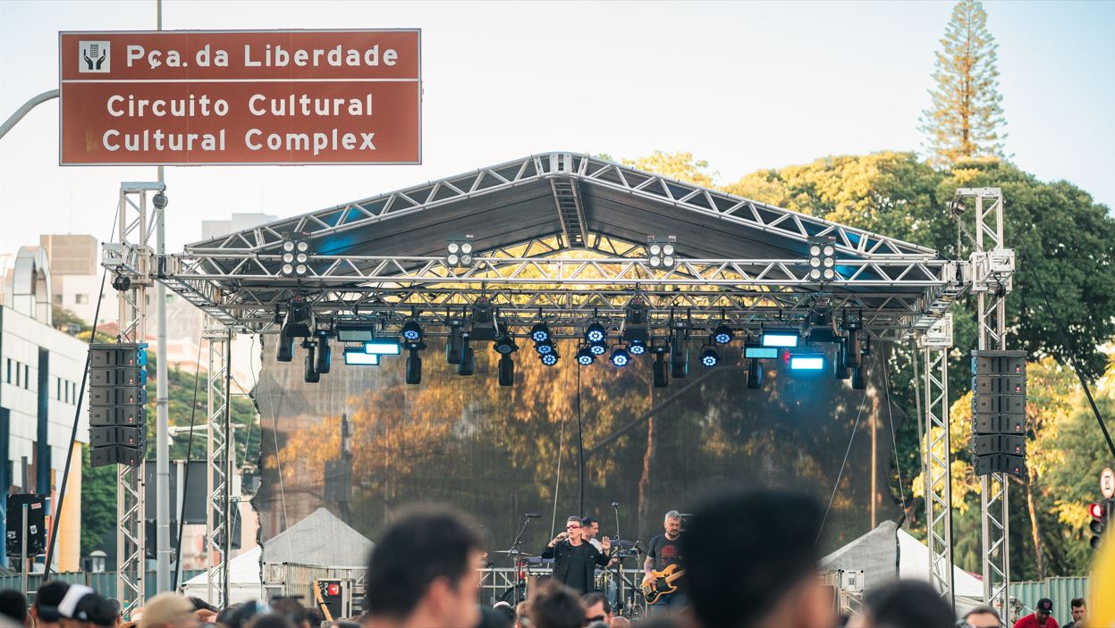 Palco montado ao ar livre na Praça da Liberdade, em Belo Horizonte, com banda se apresentando ao vivo diante de um público atento durante evento cultural.
