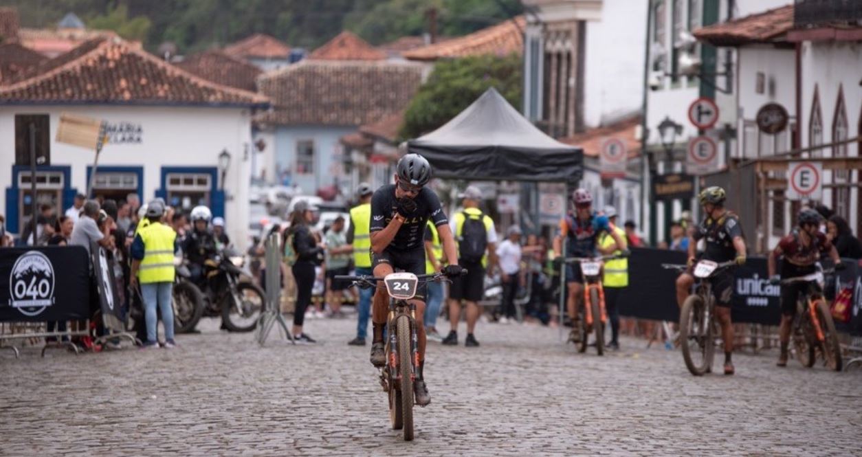 Ciclista cruza a linha de chegada na Praça Gomes Freire, em Mariana (MG), durante o Iron Biker Brasil 2025, cercado por público e construções históricas.