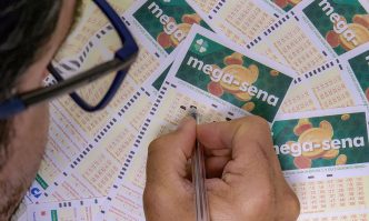 Homem preenche manualmente um bilhete da Mega-Sena, cercado por diversos volantes da loteria com o logo em destaque.