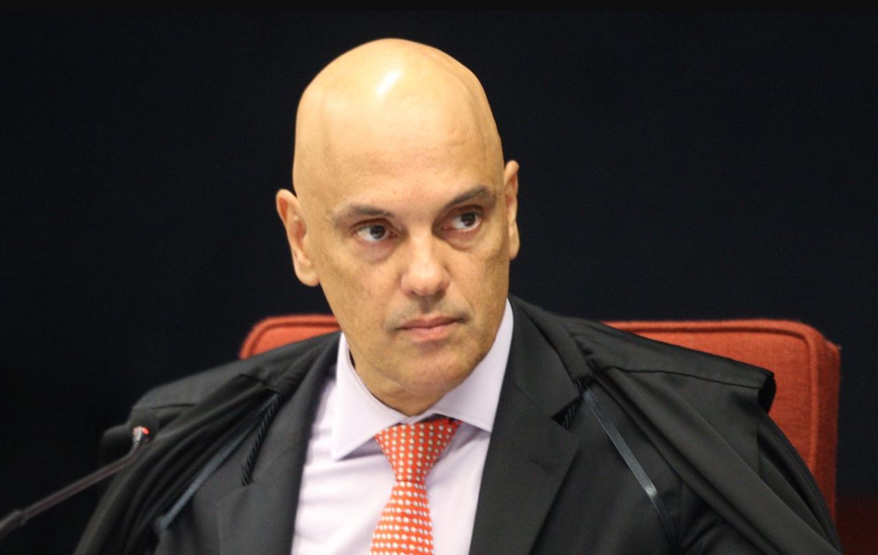 Retrato do Ministro do Supremo Tribunal Federal, Alexandre de Moraes, com expressão séria, vestindo toga preta e gravata vermelha, sentado em cadeira vermelha durante sessão oficial.