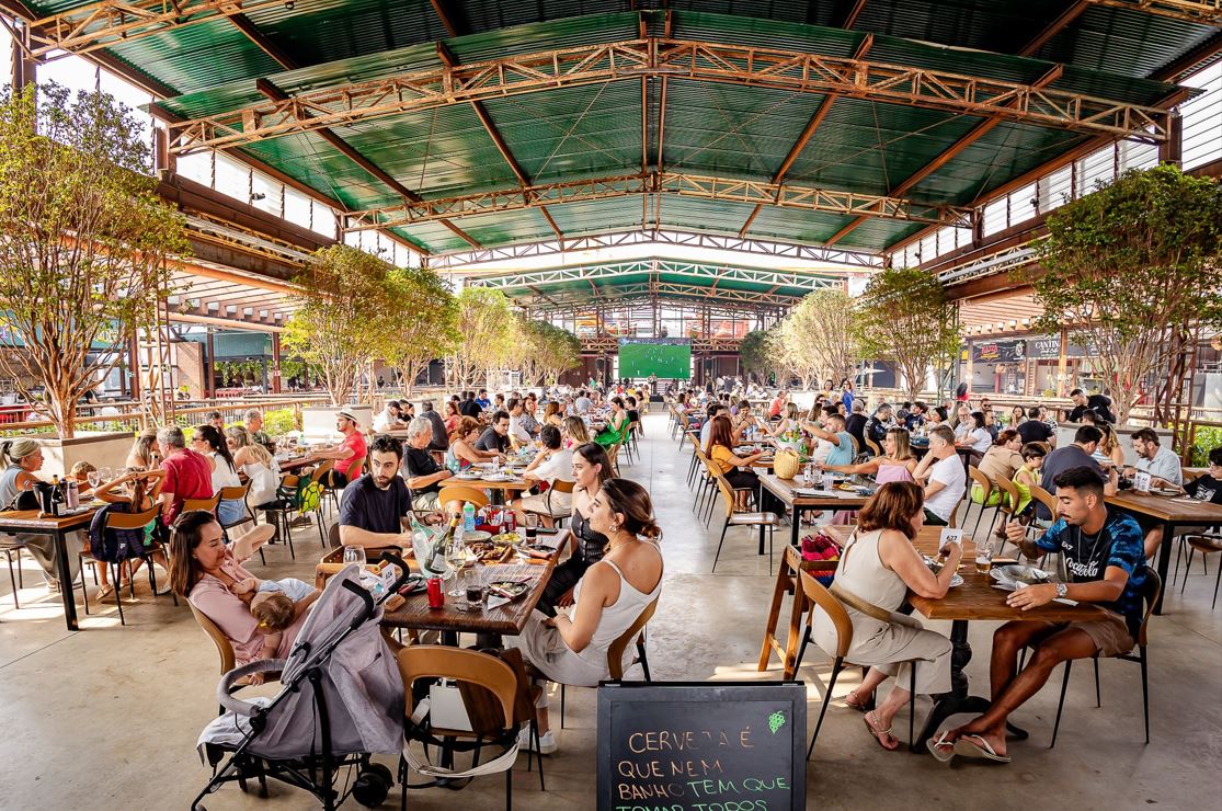 Público diverso aproveita o Festival de Pastel no Mercado de Origem, em Belo Horizonte, reunido em mesas sob galpão coberto e rodeado por árvores, em um ambiente arejado, familiar e gastronômico.