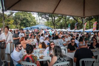 Multidão reunida sob tendas e mesas brancas na Praça José Mendes Júnior, durante evento cultural ao ar livre com clima festivo e gastronômico.