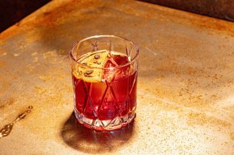 Copo de vidro lapidado com Negroni clássico, servido com gelo e fatia de laranja desidratada sobre bancada metálica rústica.