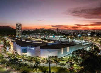 Vista aérea do BH Shopping ao entardecer, com céu alaranjado ao fundo e iluminação urbana destacando o entorno do centro comercial.