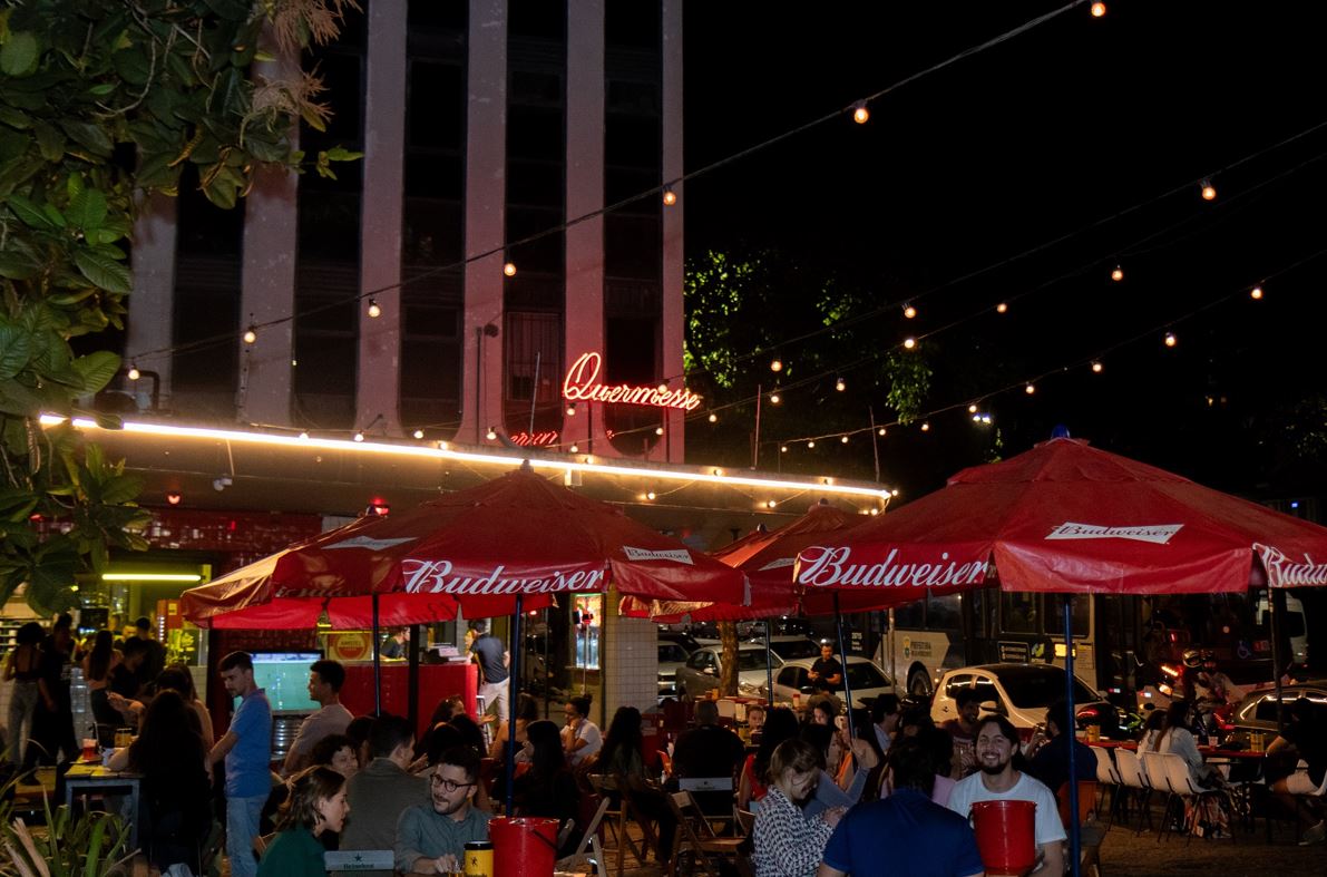Clientes aproveitam o ambiente externo do bar Quermesse em Belo Horizonte à noite, sob luzes decorativas e guarda-sóis vermelhos da Budweiser, em clima descontraído na Praça Tiradentes.