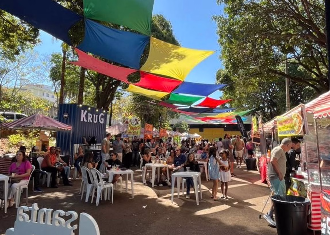 Público aproveita a Feira do Santa Tereza sob tendas coloridas, com mesas ao ar livre, barracas de comida e artesanato em ambiente arborizado.
