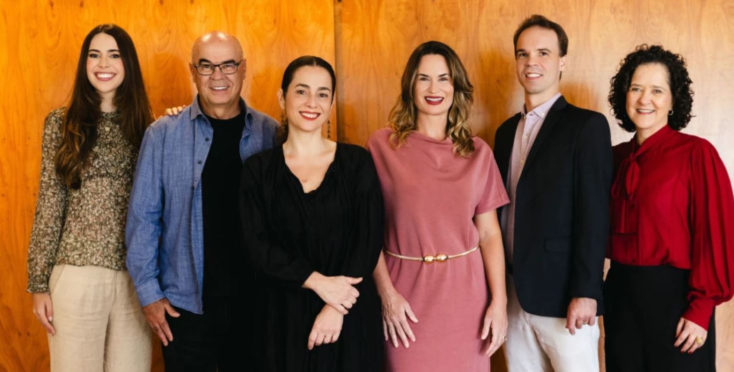 Seis pessoas posam lado a lado, sorrindo, em frente a uma parede de madeira clara. Da esquerda para a direita: Luiza Lacerda, Dino Sávio, Cris Cortez, Grasi Scalioni, Dino Bastos e Carla Madeira, integrantes da Lápis Raro e do Grupo Partners. Eles estão vestidos com roupas sociais casuais e demonstram entusiasmo pela nova fase empresarial após a aquisição.