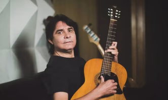 Lô Borges, músico mineiro e ícone do Clube da Esquina, segurando um violão de madeira em ambiente interno, com expressão serena.