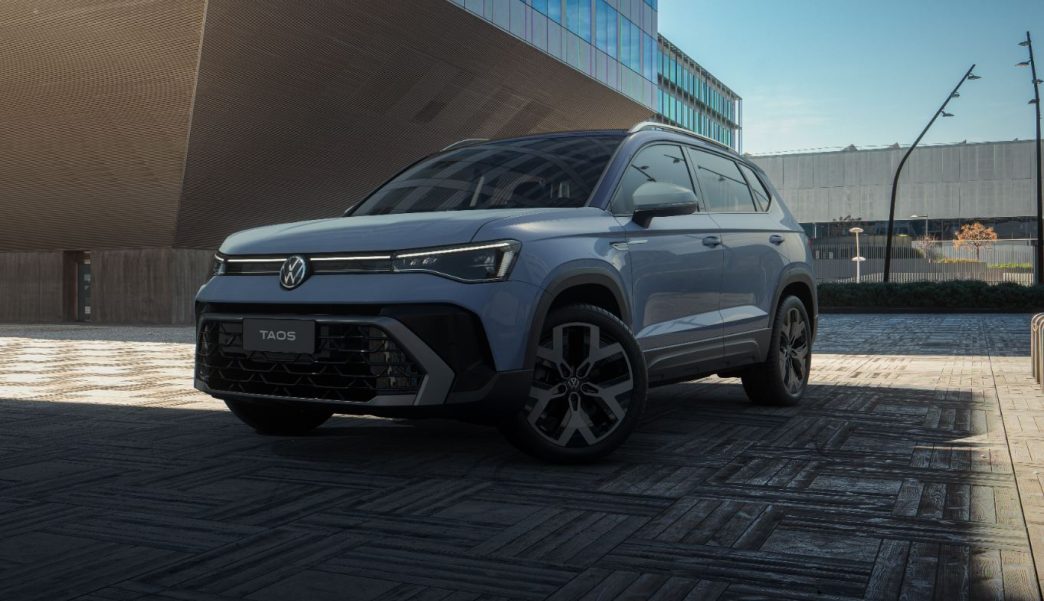 Novo Volkswagen Taos 2026 na cor Azul Pacífico estacionado em ambiente urbano moderno, destacando o novo design frontal com grade redesenhada, faróis IQ.Light em LED e rodas de liga leve aro 19 da versão Highline.