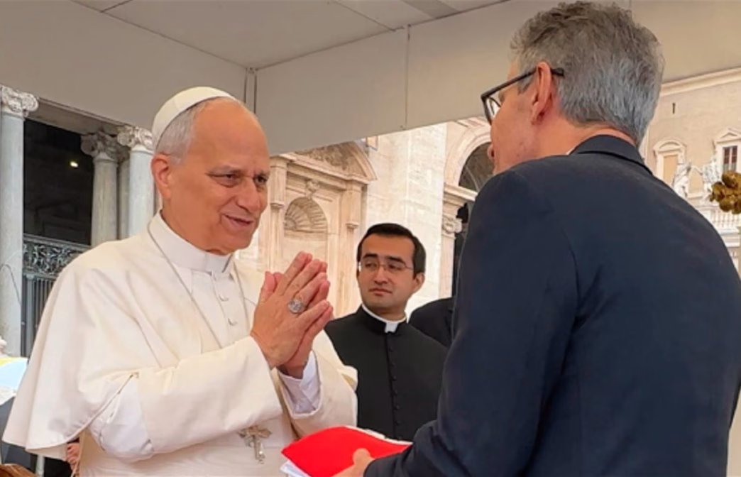 Governador Romeu Zema entrega presentes ao Papa Leão XIV durante audiência pública no Vaticano, com gesto de reverência mútua entre os líderes. A cena ocorre sob cobertura externa da Praça São Pedro, com fundo arquitetônico clássico e presença de membros do clero.