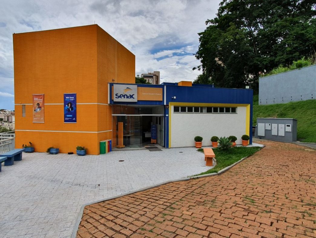 Fachada do prédio do Senac em Patos de Minas, com entrada principal, paredes em tons de laranja e azul, vegetação e bancos na área externa.