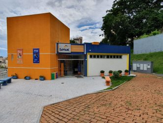 Fachada do prédio do Senac em Patos de Minas, com entrada principal, paredes em tons de laranja e azul, vegetação e bancos na área externa.
