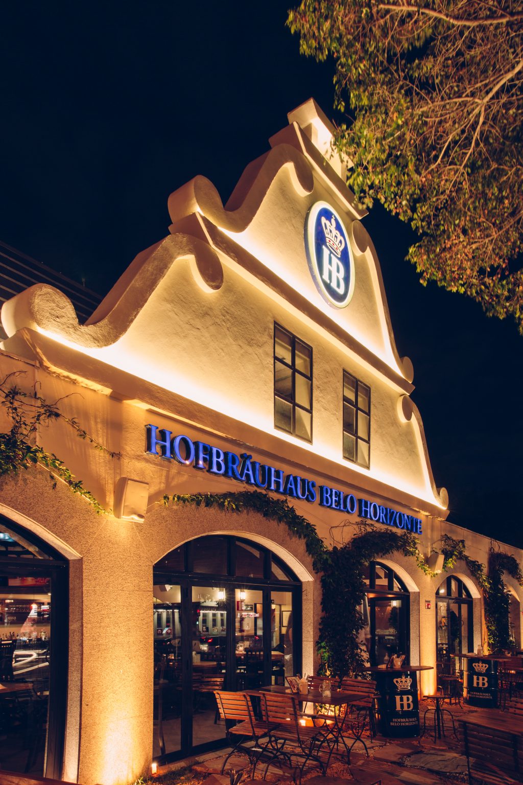 Fachada iluminada da Hofbräuhaus Belo Horizonte à noite, com arquitetura inspirada no estilo bávaro, mesas externas e letreiro em destaque.