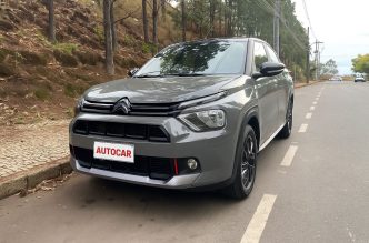 Fotografia do Citroën Basalt Dark Edition T200 estacionado em via urbana ladeada por árvores, destacando o design frontal com faróis afilados, pintura cinza Sting Gray com detalhes escurecidos e rodas pretas de liga leve.