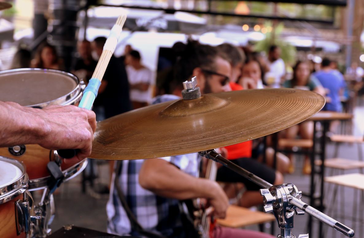 Close de um baterista tocando pratos de bateria em apresentação ao ar livre durante o evento Café com Rock no Porks Castelo, com público ao fundo.