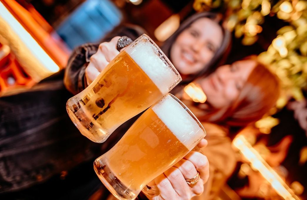 Duas mulheres brindam com copos de chope artesanal em festival gastronômico com clima descontraído e iluminação aconchegante.