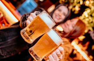 Duas mulheres brindam com copos de chope artesanal em festival gastronômico com clima descontraído e iluminação aconchegante.