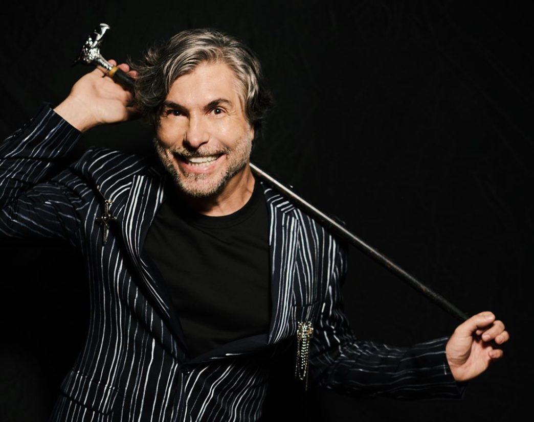 Fred Izak em pose carismática com figurino listrado e bengala artística sobre os ombros, em fundo preto, promovendo o show Lírico.