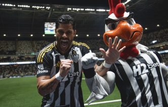 Jogador Hulk, do Atlético Mineiro, comemora intensamente ao lado do mascote Galo Doido, vestindo uniforme preto e branco, na Arena MRV lotada durante a partida contra o Bahia.