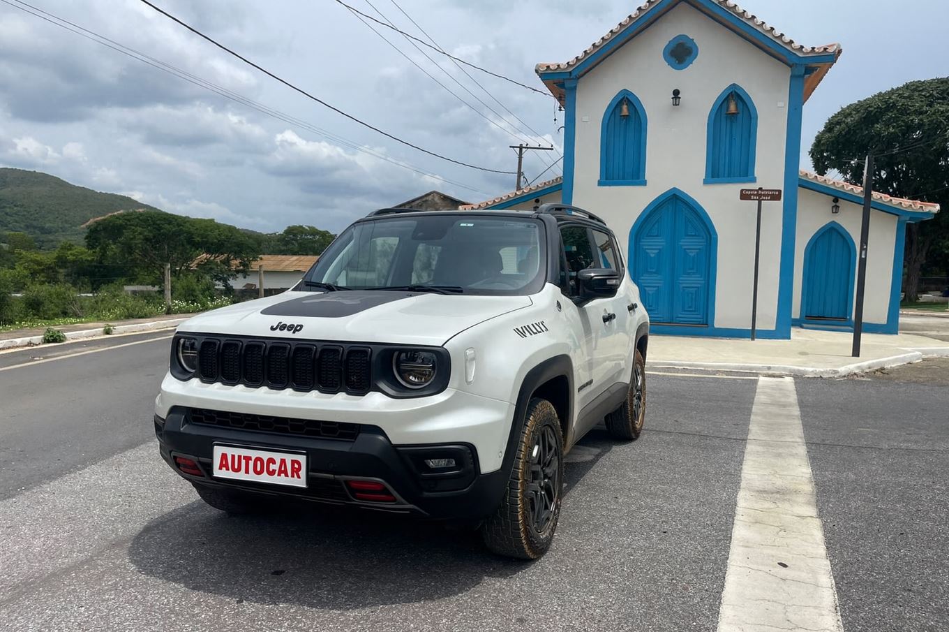 Jeep Renegade Willys 10 Anos branco estacionado em frente a uma igreja histórica com portas e janelas azuis, em estrada urbana de paralelepípedo, sob céu parcialmente nublado.