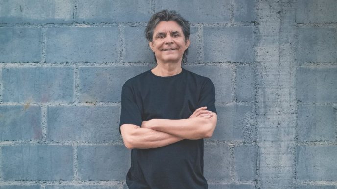 Lô Borges em pé diante de uma parede de blocos cinza, sorrindo, com os braços cruzados, vestindo camiseta preta. Foto tirada em sua fase madura.