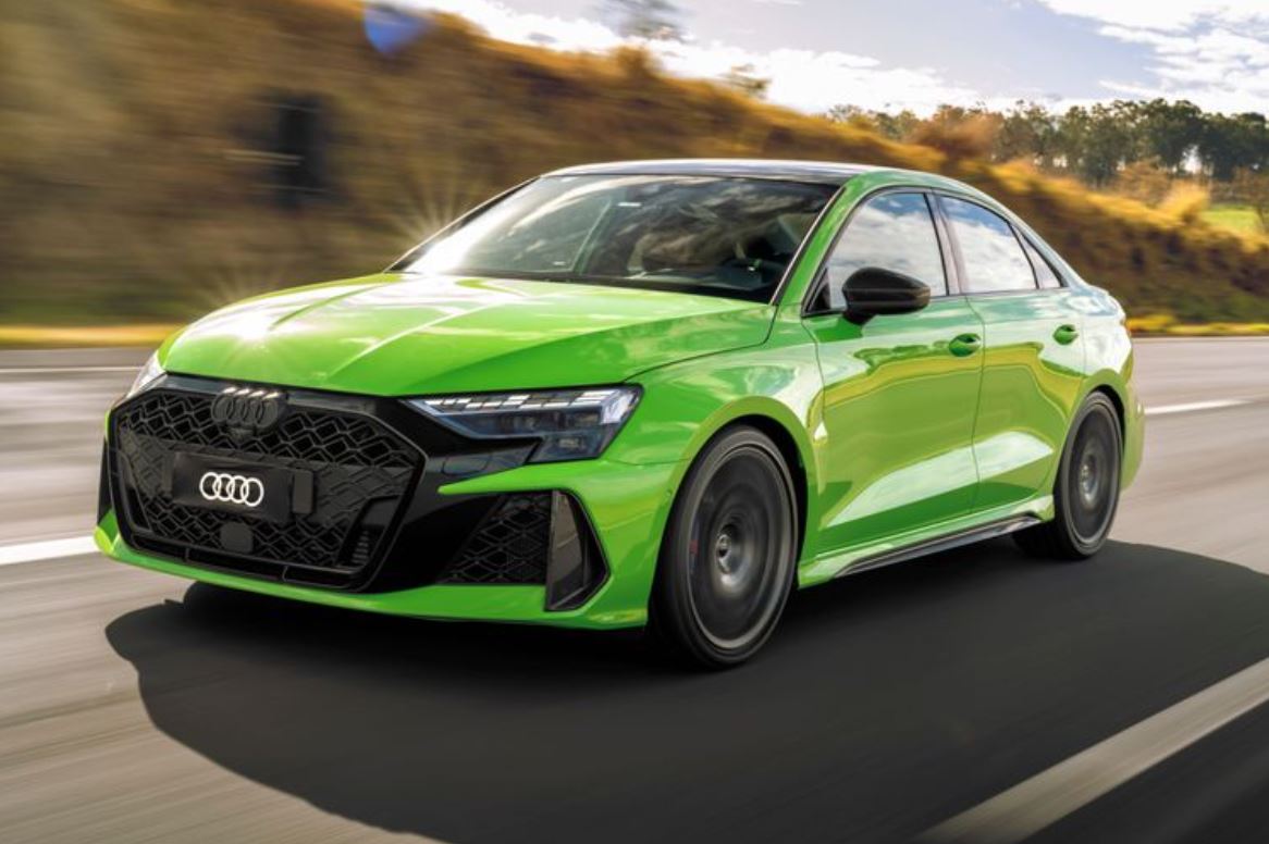 Audi RS 3 Sedan na cor Verde Kyalami em alta velocidade em rodovia aberta, destacando seu design esportivo, grade frontal agressiva e rodas de liga leve aro 19.