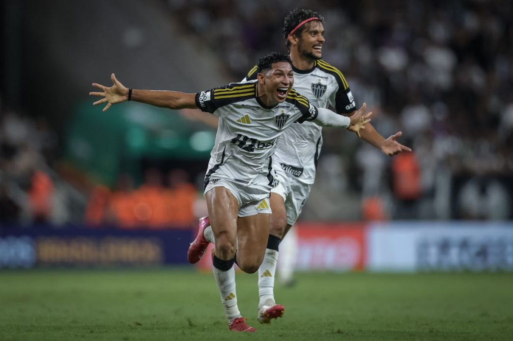 Jogador do Atlético Mineiro, vestindo uniforme branco com detalhes pretos e dourados, comemora efusivamente após marcar gol contra o Sport Recife, com os braços abertos em gesto de euforia; ao fundo, outro jogador do Galo acompanha o momento enquanto atleta do Sport permanece sentado no gramado, evidenciando o domínio alvinegro na partida válida pelo Brasileirão.