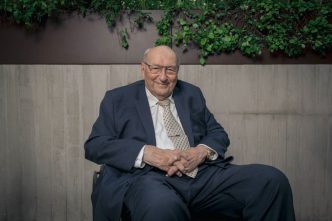 Senhor de paletó e gravata sentado em uma cadeira, com um sorriso no rosto.