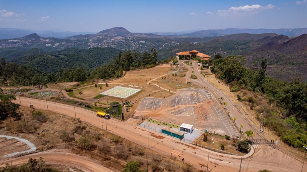 Vista aérea da antiga Mina de Águas Claras, em Nova Lima (MG), destacando o edifício histórico no topo da colina, o heliponto central e o amplo estacionamento, cercados por vegetação nativa e montanhas da Serra do Curral ao fundo.