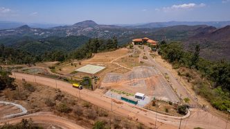 Vista aérea da antiga Mina de Águas Claras, em Nova Lima (MG), destacando o edifício histórico no topo da colina, o heliponto central e o amplo estacionamento, cercados por vegetação nativa e montanhas da Serra do Curral ao fundo.