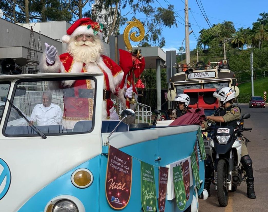 Papai Noel acena sorridente em desfile natalino a bordo de uma Kombi decorada no Natal Solidário Vila da Serra, acompanhado por policiais militares e personagens natalinos.