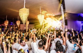 Público animado celebra a chegada do ano novo no Réveillon Porcão 2026, em ambiente sofisticado com lustres de cristal e show ao vivo, todos vestidos de branco, com taças e celulares erguidos, em clima de festa e euforia.