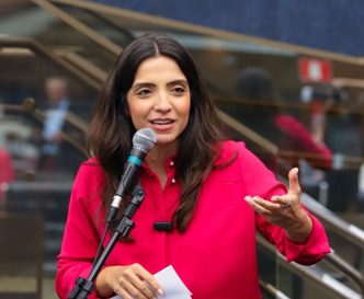 Vereadora Marcela Trópia discursa ao microfone durante apresentação pública do seu relatório de mandato, usando camisa rosa vibrante.