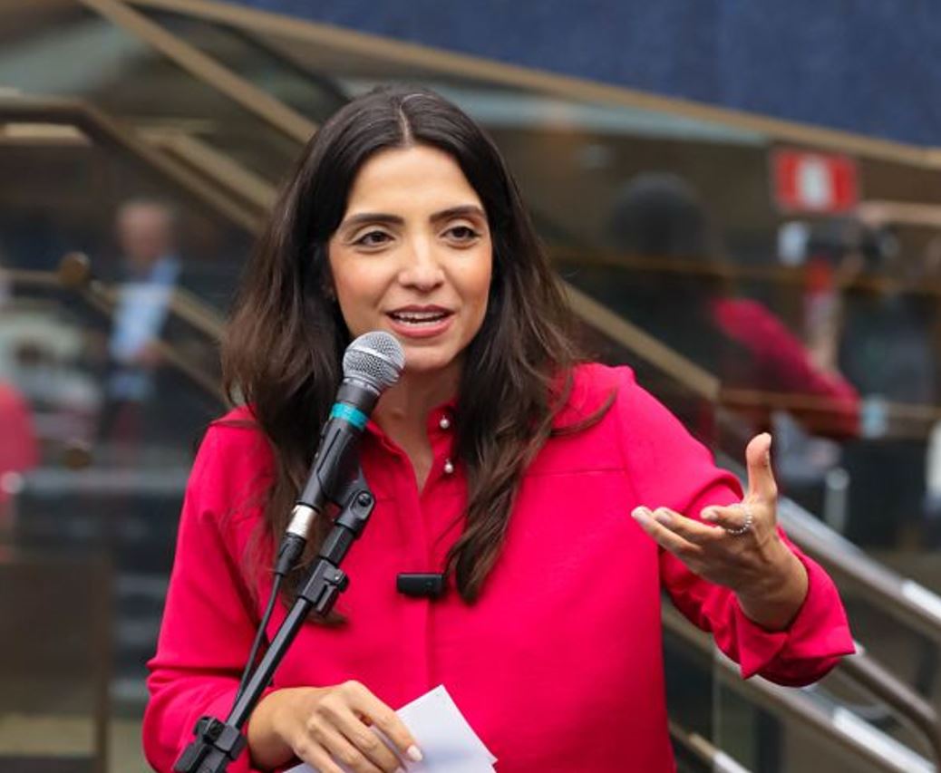 Vereadora Marcela Trópia discursa ao microfone durante apresentação pública do seu relatório de mandato, usando camisa rosa vibrante.