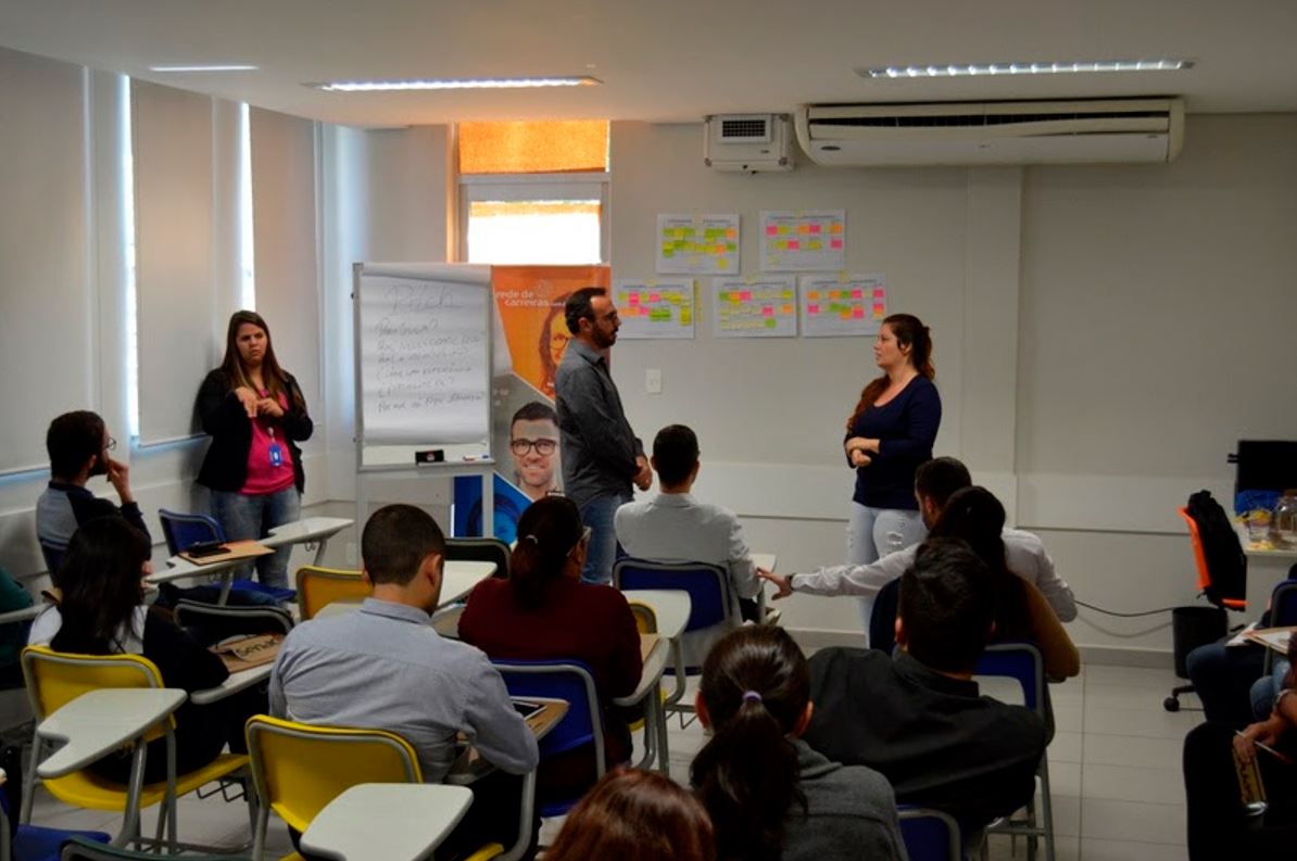Sala de aula com alunos participando de atividade prática durante curso profissionalizante; três instrutores conduzem dinâmica interativa.