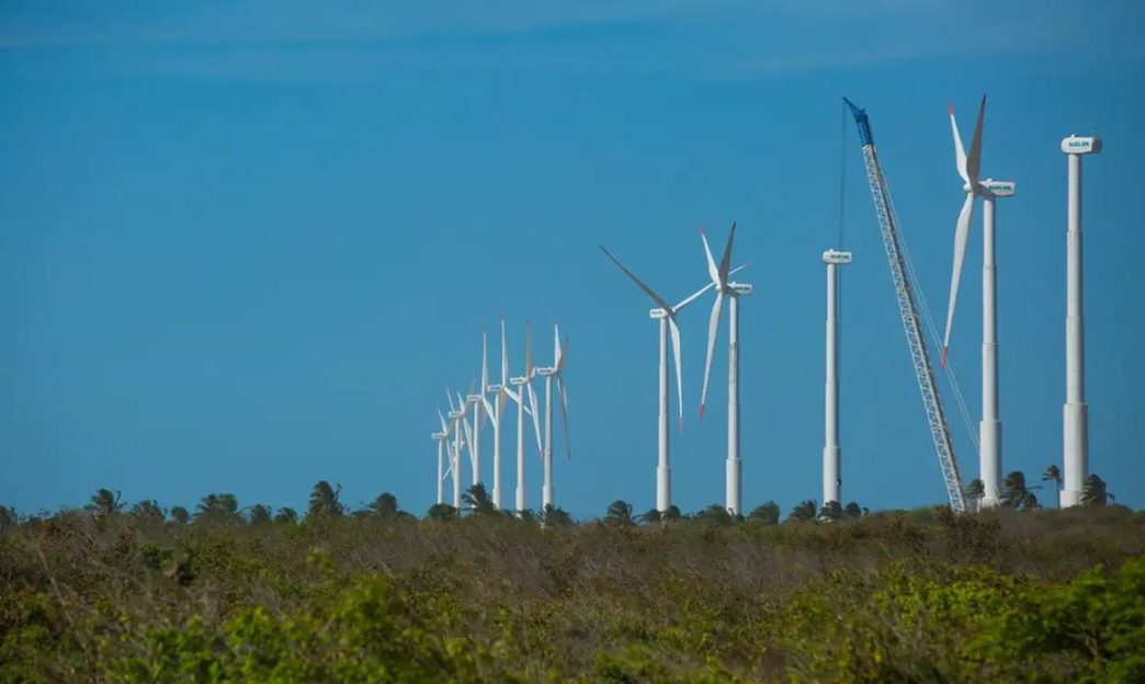 Parque eólico em construção com diversas turbinas eólicas alinhadas, cercadas por vegetação nativa e céu azul claro ao fundo.