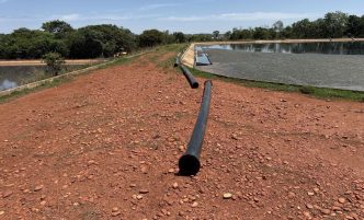 Tubulação preta de grande porte sobre solo avermelhado ao lado de lagoa da Estação de Tratamento de Esgoto da Copasa em Frutal, com vegetação ao fundo e céu parcialmente nublado.